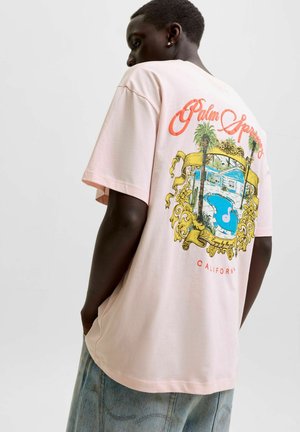 Personne portant un t-shirt rose clair avec un graphisme coloré de Palm Springs Californie montrant une piscine, des palmiers et une maison au dos.