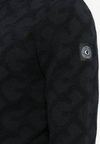 Schwarzer strukturierter Stoff mit subtilem "G"-Muster und einem kleinen runden Guess-Logo-Patch am Ärmel.