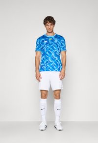 Camisola azul com padrão abstrato, mangas curtas, calções brancos, meias brancas e sapatos de futebol. Apresenta detalhes de logotipo e colarinho.