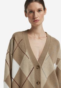 Swetrowy kardigan w kolorze beżowym z wzorem diamentu w odcieniach brązu, kremu i jasnoniebieskiego. Posiada głęboki dekolt w serek oraz dwa duże brązowe guziki.