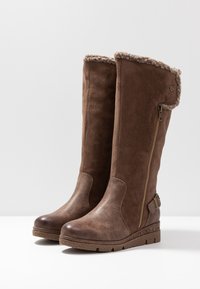 Para wysokich brązowych zimowych butów z wykończeniem z polaru, zamkiem błyskawicznym z boku, dekoracyjną klamrą i teksturowanymi gumowymi podeszwami na białym tle.