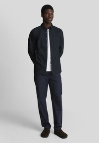 Camisa de botones azul oscuro con un único bolsillo en el pecho, llevada sobre una camiseta blanca, combinada con pantalones azul marino y zapatos marrones.