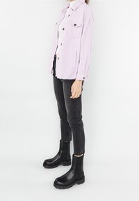 Lilažová manšestrová bunda se dvěma předními kapsami, zkombinovaná s černými úzkými džíny a černými kotníkovými botami. Na bundě je výrazný detail třásní na spodním lemu.