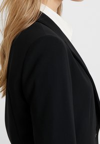 Černý blazer s hladkou texturou, strukturovanými rameny, zapínáním na jeden knoflík a klopovými límci, doplněný bílou košilí s límečkem.