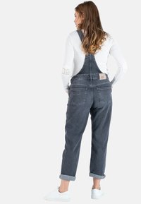 Donkergrijze denim overalls met verstelbare bandjes, voorzakken en opgerolde enkels. Gedragen over een wit shirt met lange mouwen en witte schoenen.