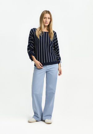 Junge Frau steht und trägt ein dunkelblaues Langarmshirt mit weißen vertikalen Streifen, hellblaue weite Hosen und weiße Sneaker.