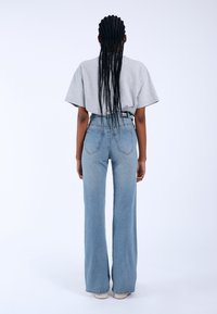 Personne vue de dos, aux longs cheveux tressés, portant un t-shirt gris ample, un jean évasé bleu taille haute et des baskets blanches.