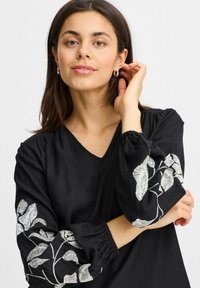 Blusa nera con scollatura a V e maniche lunghe e gonfie, con intricato ricamo bianco a forma di foglia sui polsini. Tessuto liscio, vestibilità comoda.