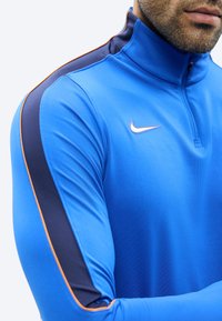 Mees kannab sinist pika varrukaga sportlikku särk, millel on ees lukk ja must triip oranži äärega varrukatel.