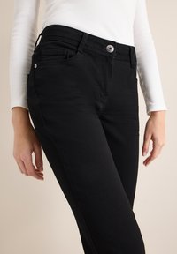 Pantalones vaqueros negros de corte slim, con cierre frontal de botones, cinco bolsillos y detalles sutiles de costura. Textura suave y lisa.