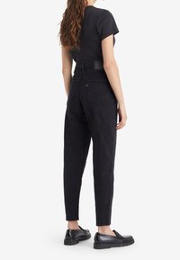 Jean noir taille haute avec une jambe fuselée, design à cinq poches et tissu texturé, associés à des chaussures plates noires.