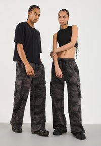 Two people standing side by side, wearing black and gray camouflage cargo pants with black tops and black chunky shoes, on a plain background.