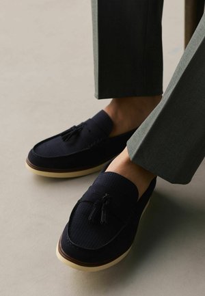 Men's dark blue suede loafers with tassels worn with gray trousers on a light floor.
