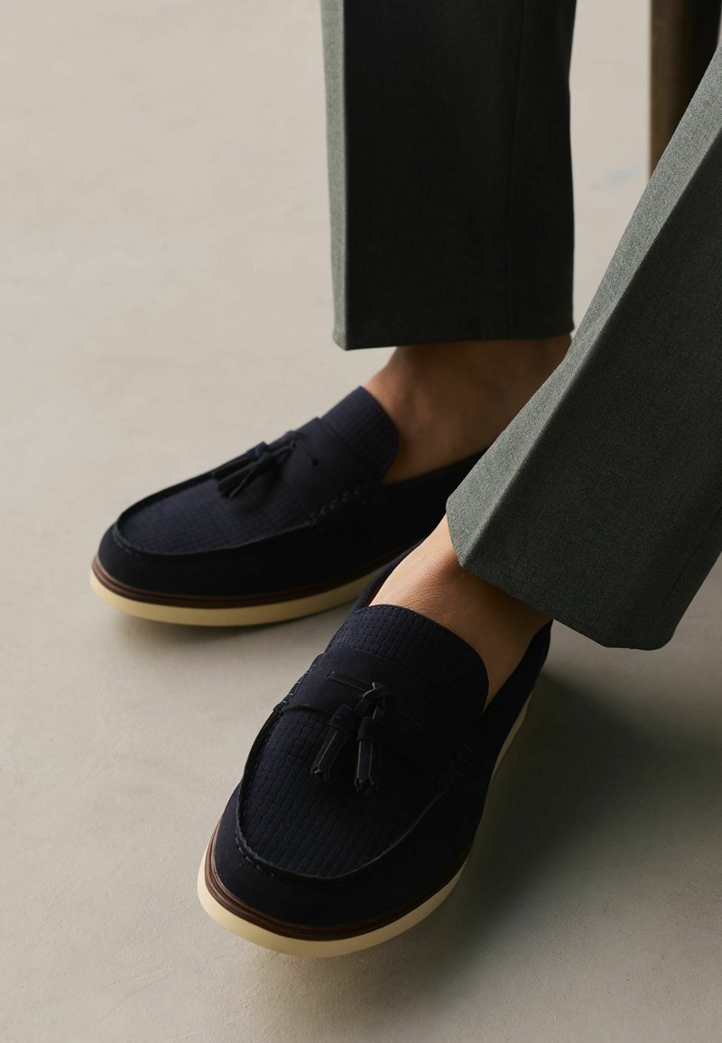 Mocassins en daim bleu foncé pour hommes avec des pampilles, portés avec un pantalon gris sur un sol clair.