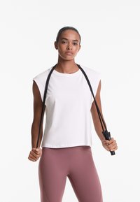 White sleeveless cotton top with a relaxed fit, paired with pink leggings. The model holds a black jump rope with ergonomic grips.
