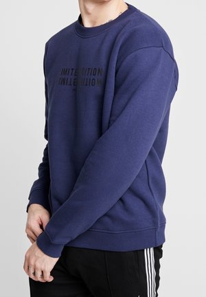 Homme portant un sweat-shirt bleu avec le texte « LIMITED EDITION » et un pantalon noir avec des bandes blanches sur les côtés sur fond gris.