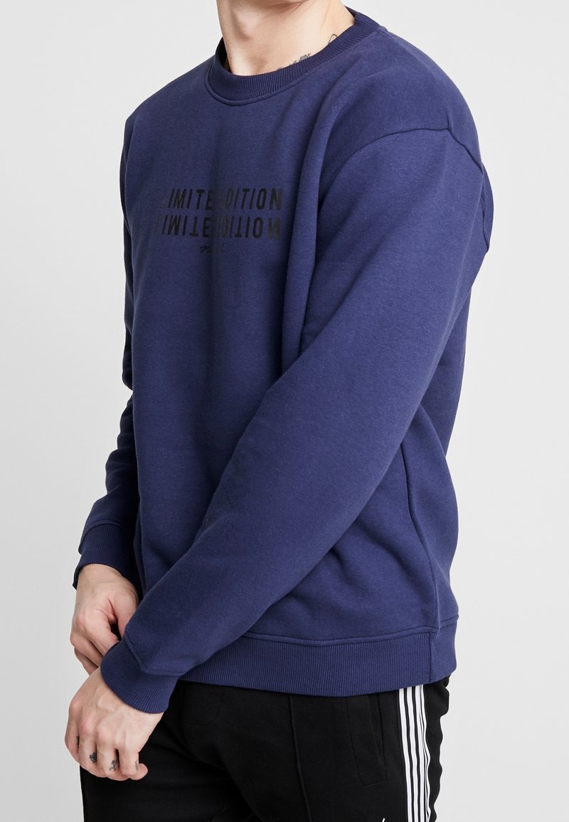 Homme portant un sweat-shirt bleu avec le texte « LIMITED EDITION » et un pantalon noir avec des bandes blanches sur les côtés sur fond gris.