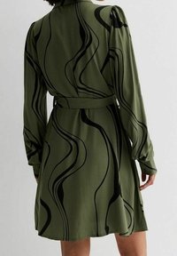 Vestido verde de manga larga, con patrones abstractos en negro que imitan olas. Cinturones en la cintura, tela suave, dobladillo acampanado y cuello redondeado.