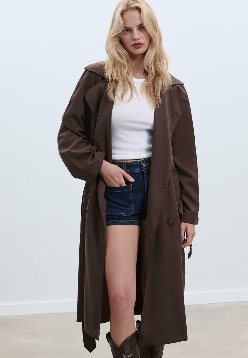Bruine trenchcoat met een ontspannen pasvorm, voorzien van grote revers, knoopsluiting en mouwen met opgerolde manchetten, gedragen over een witte top en denim shorts.