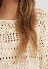 Neulepaita vaaleassa kremessä, jossa on avoin kuvio, jossa on vaihteleva reikiä ja teksturoituja raitoja, pyöreä kaula-aukko ja rento istuvuus.