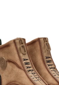 Botas de tobillo de ante marrón con cremalleras laterales, que presentan un parche con el logo y costuras en contraste. Superficie texturizada y detalle de impresión geométrica.
