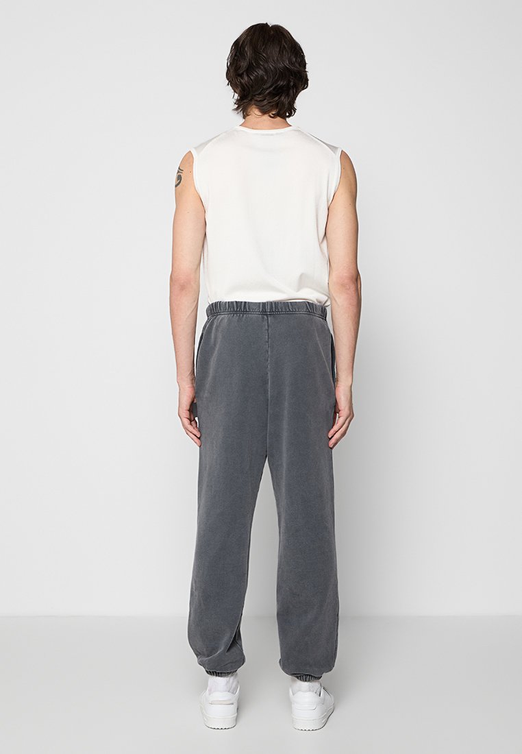 Pantalones de felpa grises, suaves, con una cintura elástica y tobillos ajustados, combinados con una blusa sin mangas de color crema. Tatuaje visible en el brazo.