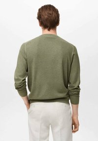 Olive green knitted sweater with a round neckline and ribbed cuffs, worn over white trousers, featuring a textured pattern.
