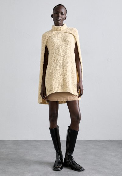 Beige textured cape with a high collar, worn over a short beige skirt. Paired with knee-high black glossy boots. Simple and modern design.