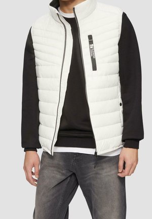 Homme portant une veste sans manches blanche matelassée sur un sweat-shirt noir et un jean noir délavé, les mains détendues le long du corps.