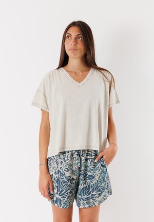 T-shirt beige chiaro a V con maniche corte abbinato a pantaloni corti blu a fantasia con un design a foglie. Il tessuto sembra morbido e traspirante.