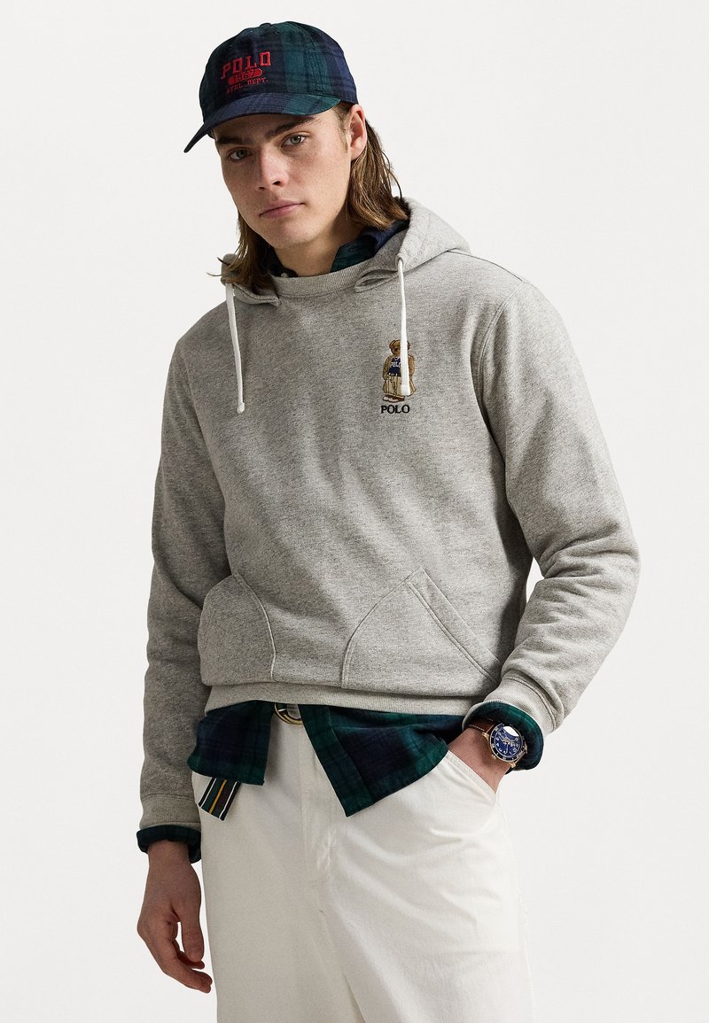 Haut en gris avec cordons blancs et poches avant. Présente un logo "Polo" brodé. Porté par-dessus une chemise à carreaux bleue et verte, associé à un pantalon blanc.