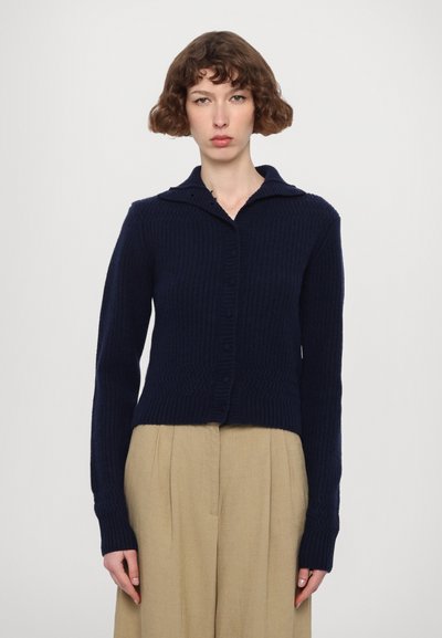 Marineblaue Strickjacke mit geripptem Muster, Knopfverschluss und spitzem Kragen. Kombiniert mit beigen, hoch taillierten Hosen.