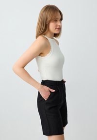 Body bianco a coste con bordi ondulati, abbinato a pantaloni corti di lino neri con tasche laterali e vestibilità sartoriale. Modello in posa di profilo.