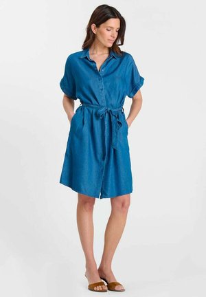 Femme portant une robe bleue boutonnée jusqu'au genou avec des manches retroussées et une taille nouée, debout les mains dans les poches, regardant vers le bas.