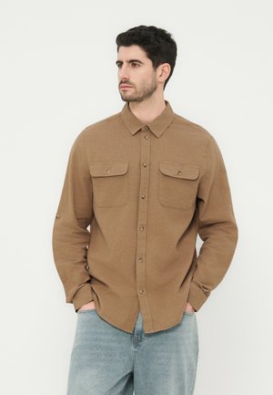 Άνδρας που φοράει καφέ πουκάμισο με κουμπιά και τσέπες με καπάκια, με ανοιχτόχρωμα μπλε τζιν, τα χέρια στις τσέπες, κοιτάζοντας στο πλάι σε λευκό φόντο.