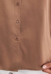 Bruine, gladde stof blouse met een doorlopende knoopsluiting, met twee knopen en een rechte zoom. Handen langs de onderrand.