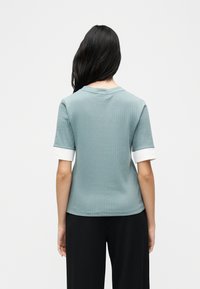 Γυναίκα με μακριά σκουρόχρωμα μαλλιά, φορώντας ανοιχτό μπλε ριμπ κοντομάνικο πάνω μέρος με λευκές μανσέτες και μαύρο παντελόνι, γυρισμένη προς τα πίσω σε λευκό φόντο.