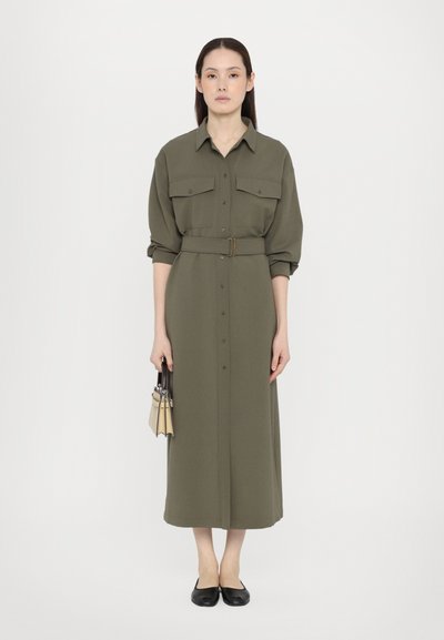 Olivgrünes langes Hemdkleid mit Knopfverschluss, zwei Brusttaschen und einem Gürtel. Getragen mit schwarzen Ballerinas und einer kleinen Handtasche.