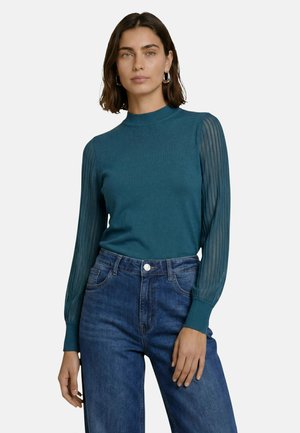 Vrouw met kort bruin haar, gekleed in een groenblauwe top met lange mouwen met doorschijnende strepen en een hoge taille blauwe spijkerbroek, staand tegen een witte achtergrond.