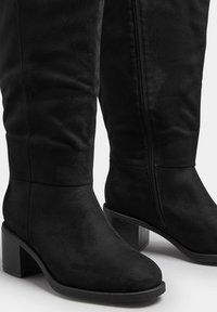 Botas de ante negras hasta la rodilla con punta cuadrada y tacón ancho. Cuenta con una cremallera lateral para facilitar su uso. Superficie texturizada y diseño minimalista.