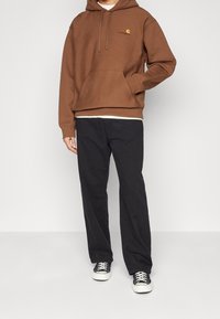Felpa cappuccio marrone con tasca frontale e logo sul petto, abbinata a pantaloni neri a gamba dritta e sneakers con motivo.