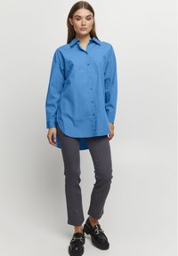 Camicia azzurra in cotone con colletto a punta, maniche lunghe e orlo arrotondato, abbinata a pantaloni grigi slim-fit e mocassini neri.
