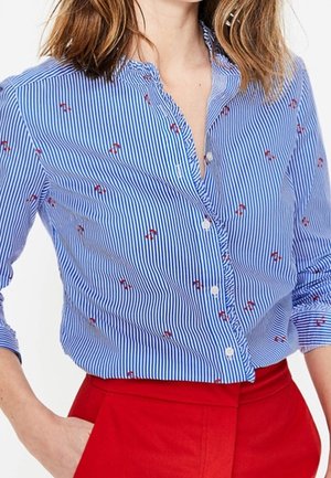 Woman wearing a blue and white striped shirt with small red flamingos, tucked into high-waisted bright red pants, with one hand in pocket.