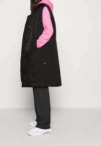 Svart ärmlös jacka med tryckknappar, bärs över en rosa huvtröja, kombinerad med gråbyxor och vita sneakers.