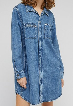 Kobieta w niebieskiej sukience koszulowej z denimu z długimi rękawami, kieszeniami na piersi — w tym jedna z zamkiem błyskawicznym — oraz kołnierzykiem.