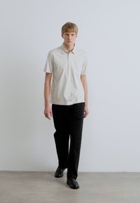 Béžové polokošeľa s krátkym rukávom a tromi gombíkmi, doplnená čiernymi nohavicami s rovnými nohavicami. Oblečenie dopĺňajú čierne topánky.