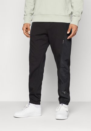 Jogging noir en tissu technique avec un panneau texturé et une coupe slim, associé à des baskets blanches. Le matériau semble doux et extensible.