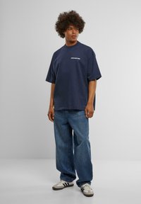 Marineblå oversized t-shirt med teksten "FORBUDT", lavet af bomuldsmateriale, med korte ærmer, parret med lyseblå baggy jeans og sneakers.