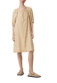 Abito in lino beige con scollo quadrato e maniche a sbuffo, silhouette fluida, abbinato a sandali beige con cinturini su uno sfondo neutro.