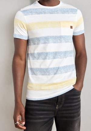 Homme portant un T-shirt blanc à manches courtes avec des rayures horizontales bleu et jaune délavées et une poche poitrine, assorti à un jean noir.